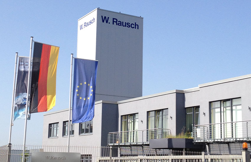 Außenfassade der Firma W. Rausch mit einer Europflagge, Deutschlandflagge und Firmenflagge mit dem Dino im Vordergrund